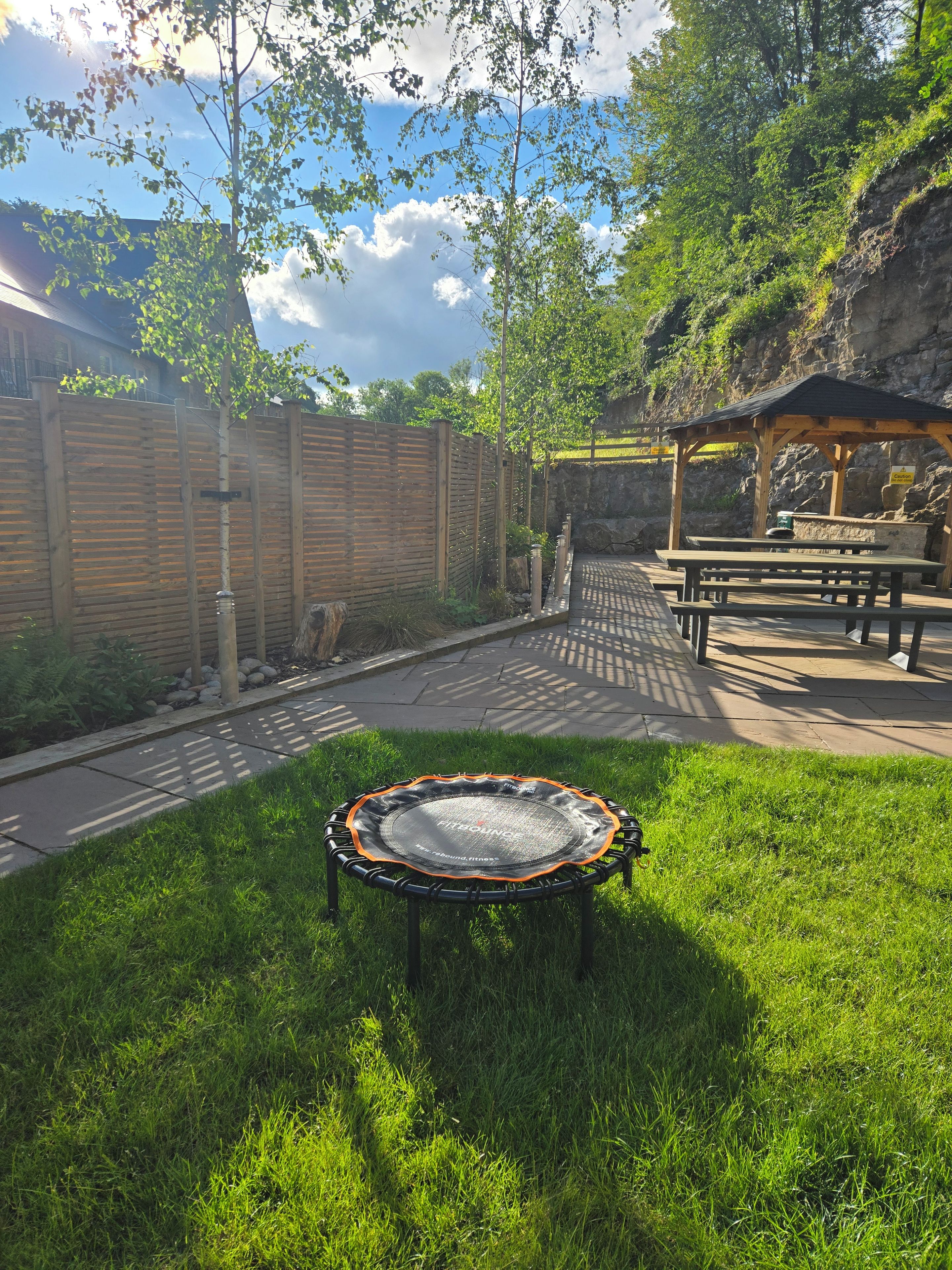 A trampoline in a grassy area with picnic tables and a wooden structure in the background.