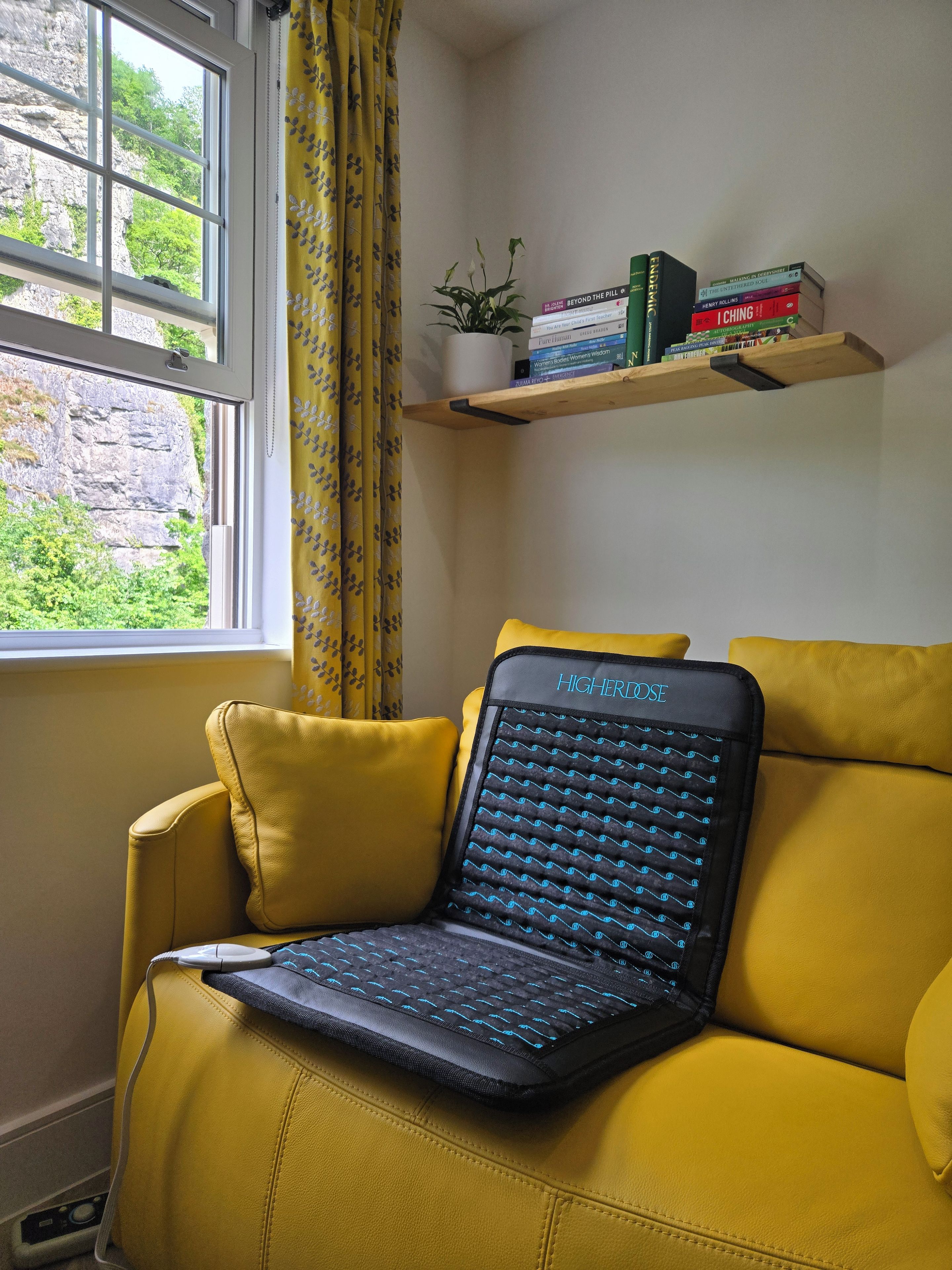 Un sofá amarillo con una computadora portátil y un cojín, una ventana con cortinas, y una estantería con libros.