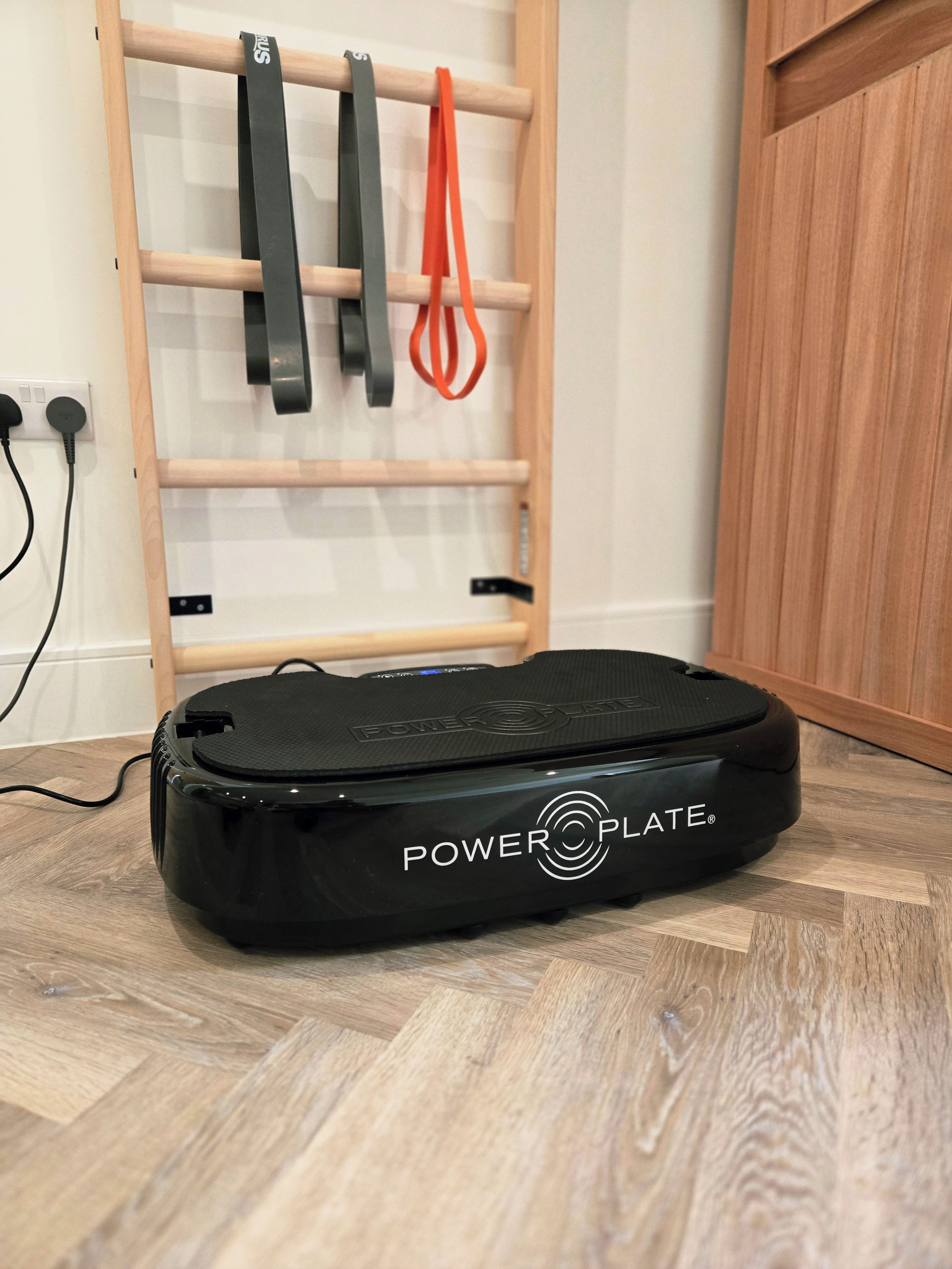 A PowerPlate exercise machine in a room with a wooden floor and a wall-mounted rack with resistance bands.