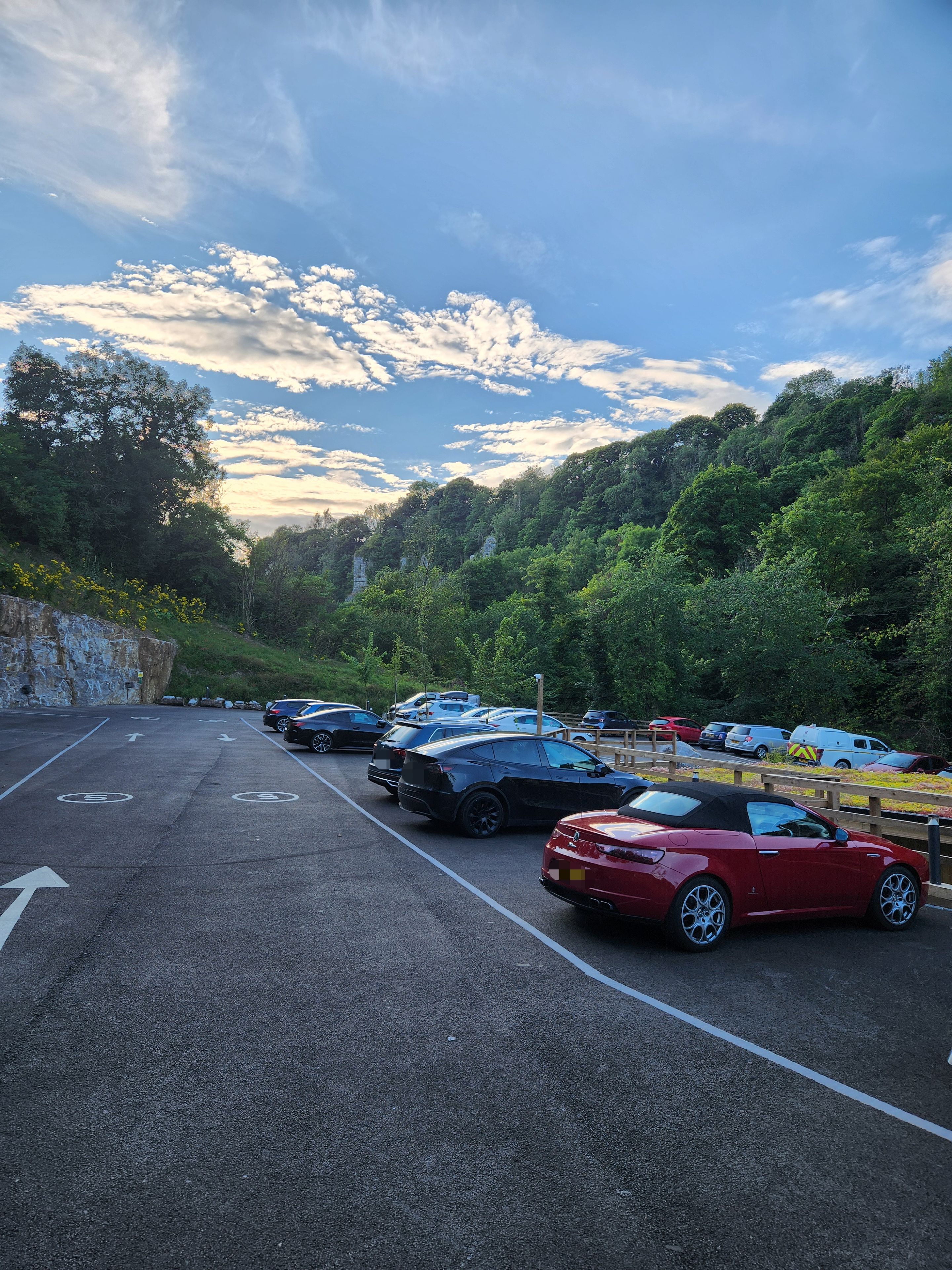 A parking lot with multiple cars, a stone wall, and a forested hillside in the background.