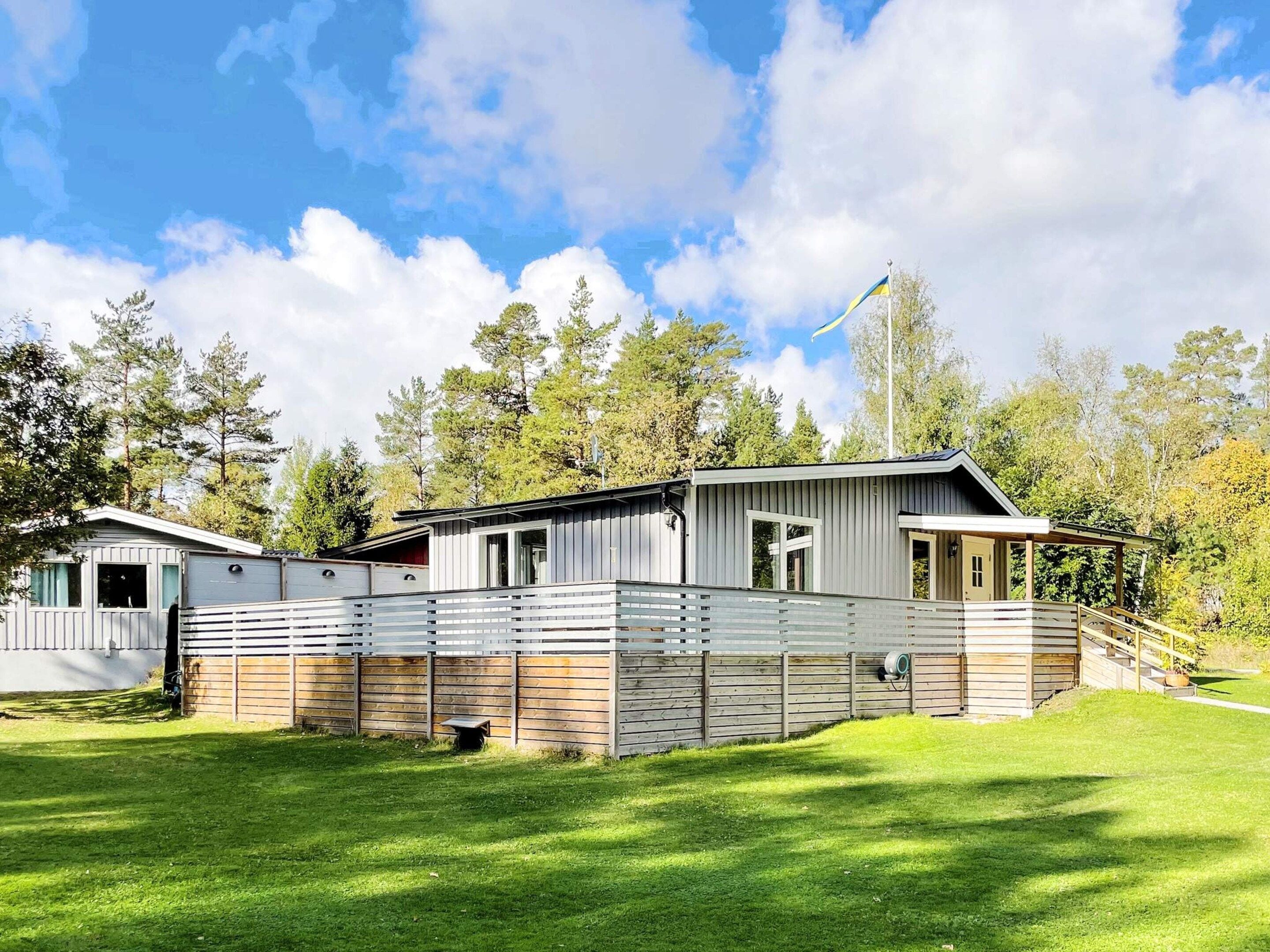 Uma casa móvel moderna no estilo de trailer com um quintal cercado, rodeada por árvores e um céu claro.