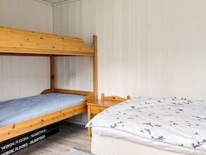Ein Stockbett mit einem unterem Bett und einem Einzelbett, beide mit Holzrahmen. Neben dem Einzelbett steht ein kleiner Nachttisch aus Holz.