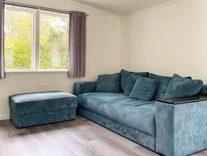 Ein Zimmer mit einer Couch, einem Hocker, einem Fenster mit Vorhängen und Blick auf Bäume.