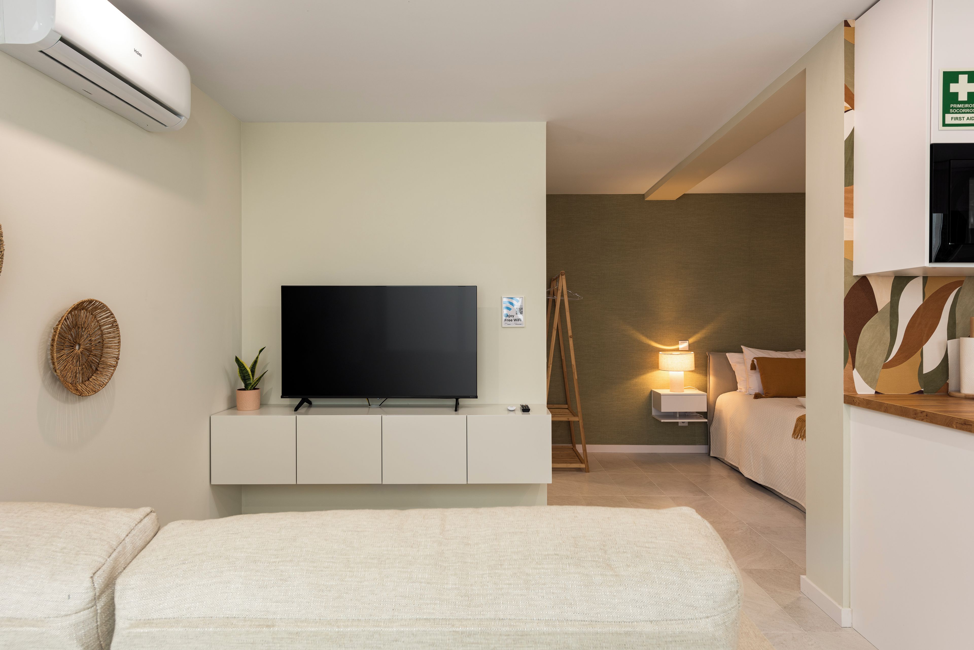 A modern hotel room with a flat-screen TV on a white cabinet, a bedside table with a lamp, a wooden ladder, and a bed with a white duvet.