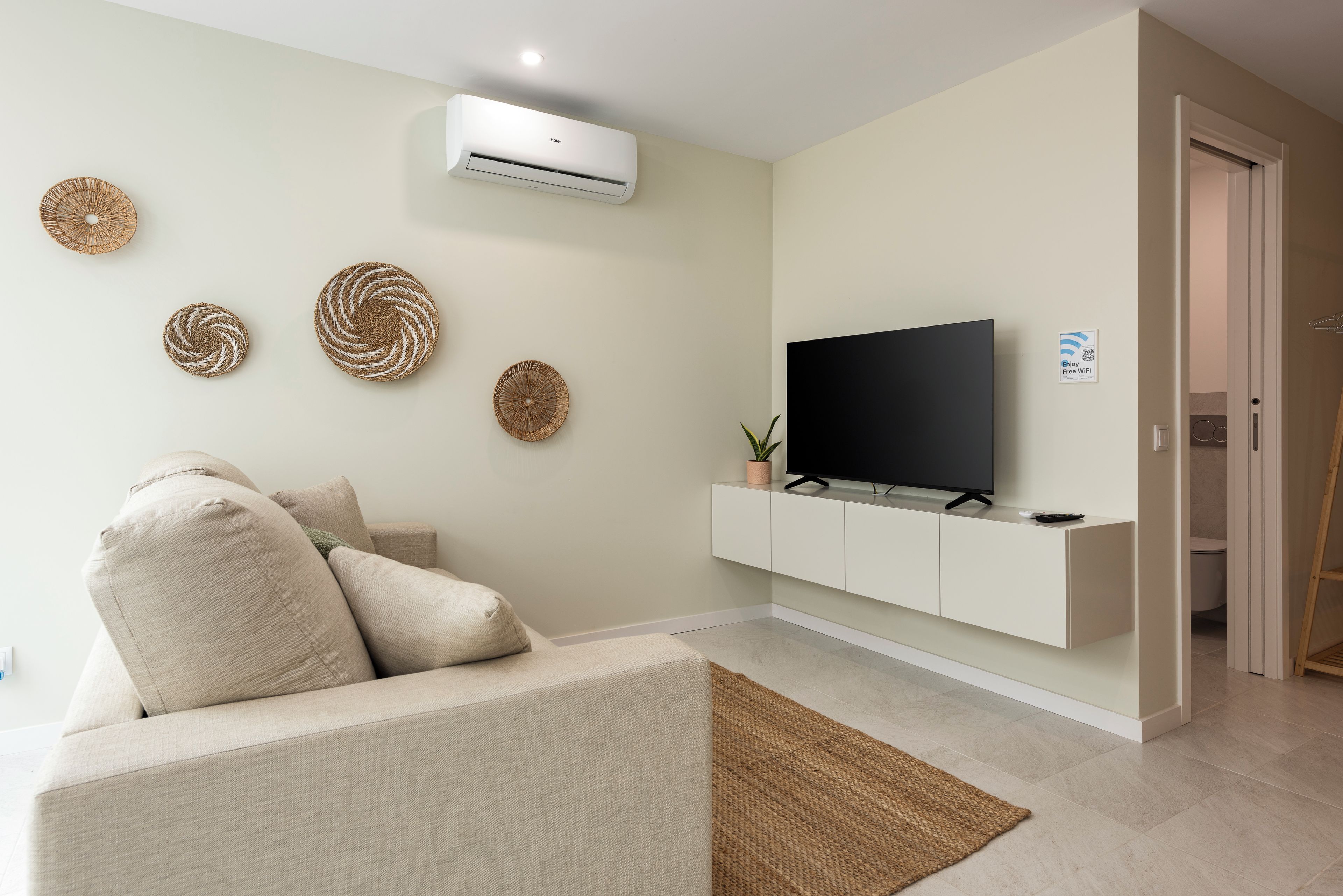 A living room with a sofa, a flat-screen TV, a mounted air conditioner, and decorative woven wall art.