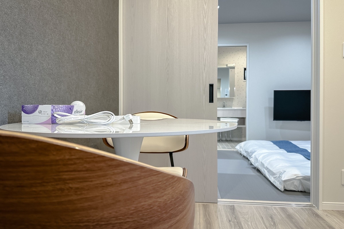 Ein Hotelzimmer mit rundem Tisch, einem Sessel, einem Bett, einem Fernseher und einem sichtbaren Badezimmer im Hintergrund.