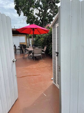 Ein überdachter Patio-Bereich mit Tisch und Stühlen, einem roten Sonnenschirm und einem weißen Gebäude im Hintergrund.