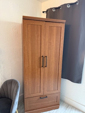 Ein hölzerner Kleiderschrank mit zwei Türen und einer Schublade, neben einem grauen Sessel und einem dunklen Vorhang.