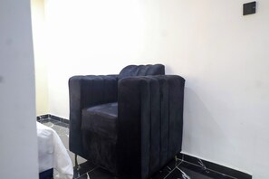 Una butaca negra tapizada en un rincón de una habitación con paredes blancas y suelo de baldosas negras.