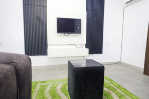 Ein modernes Wohnzimmer mit einem flachbildfernseher, der an einer weißen Unterhaltungseinheit befestigt ist, einem schwarzen Hocker in der Mitte und einem grün-weißen Teppich auf dem Boden.