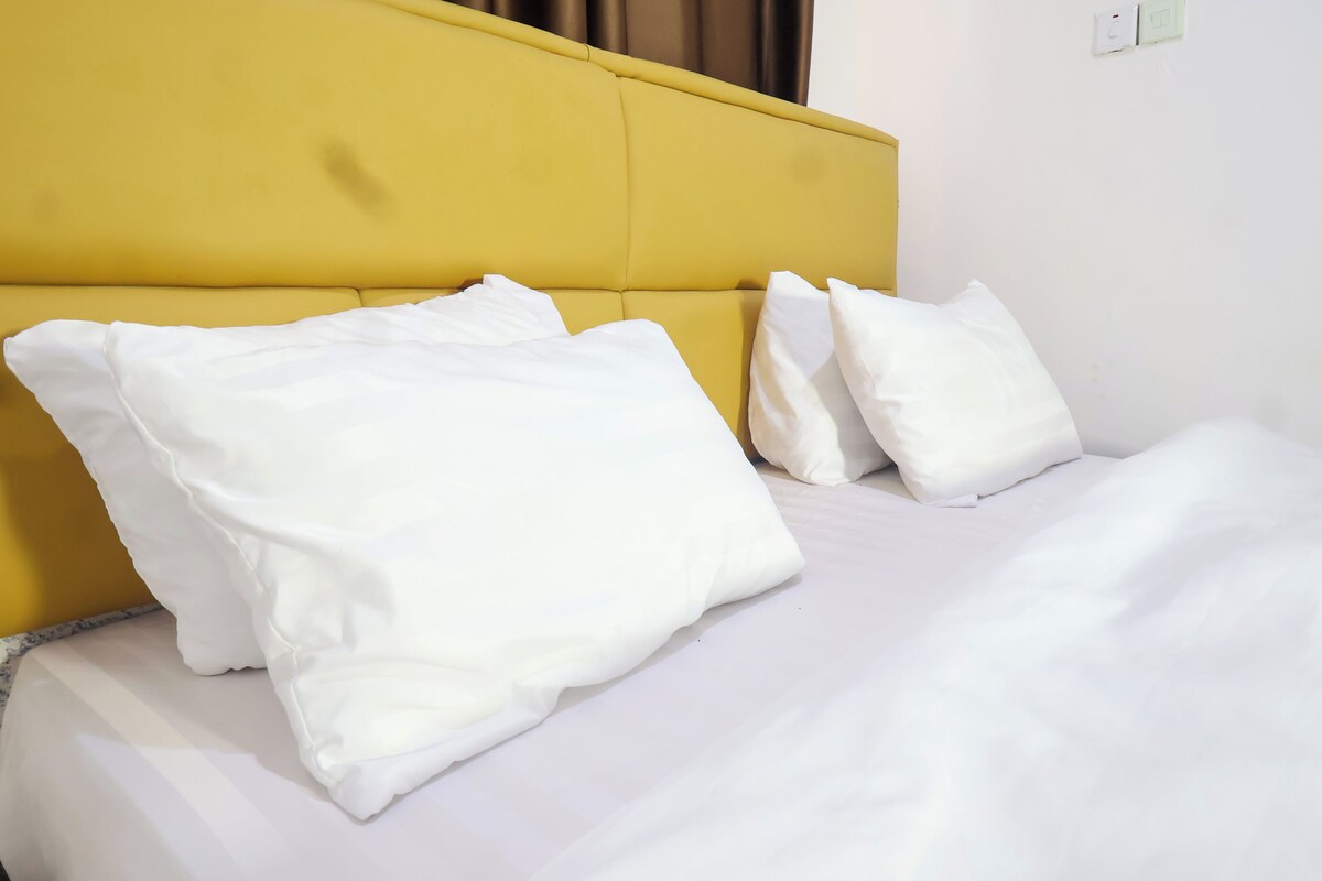 Ein Bett mit weißer Bettwäsche und Kissen, ein gelber Kopfteil und eine schlichte Wand mit Lichtschalter.