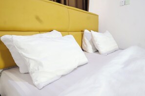 Ein Bett mit weißer Bettwäsche und Kissen, ein gelber Kopfteil und eine schlichte Wand mit Lichtschalter.