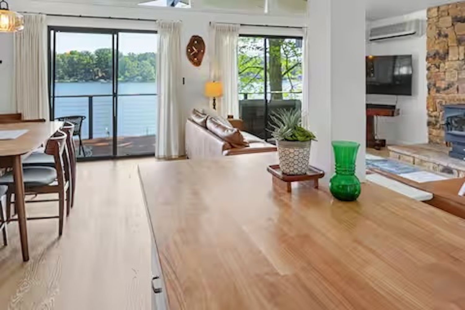 A spacious living area with a wooden dining table, a sofa, and a view of a lake through large windows.