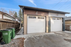 A garage with two white doors, a wooden fence, and green trash bins. - 3BR Spacious Home • Mins to UofA, Whyte & Downtown (Edmonton)