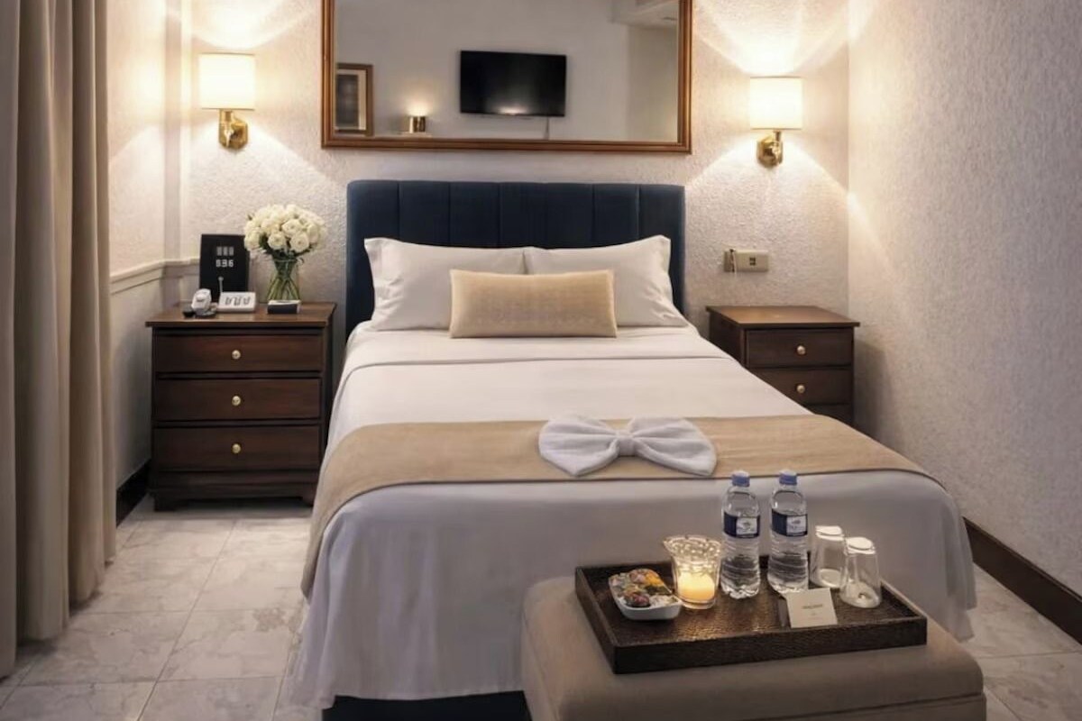 Habitación de hotel con una cama grande, televisor montado encima, lámparas de noche, mesita de noche con un reloj y flores, y una bandeja con artículos de aseo.