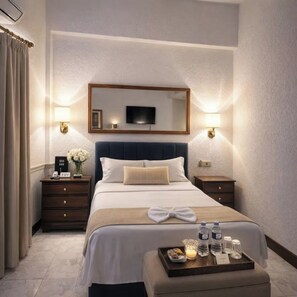 Habitación de hotel con una cama grande, televisor montado encima, lámparas de noche, mesita de noche con un reloj y flores, y una bandeja con artículos de aseo.