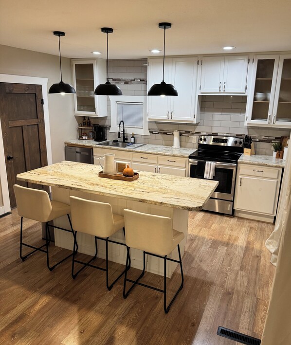 A modern kitchen with a large island, pendant lights, and wooden flooring.