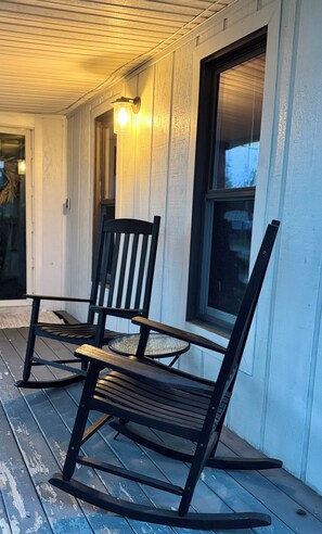 A porch with two rocking chairs and a table, wooden siding walls, and a window with blinds.