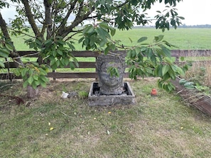 A garden with a wooden fence, a tree, and a stone planter with a statue. - Knus chalet met privé jacuzzi tussen Hoorn en Enkhuizen (Wijdenes)