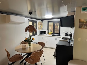 Una cocina compacta con muebles blancos, microondas y un comedor con sillas de mimbre y una mesa redonda.