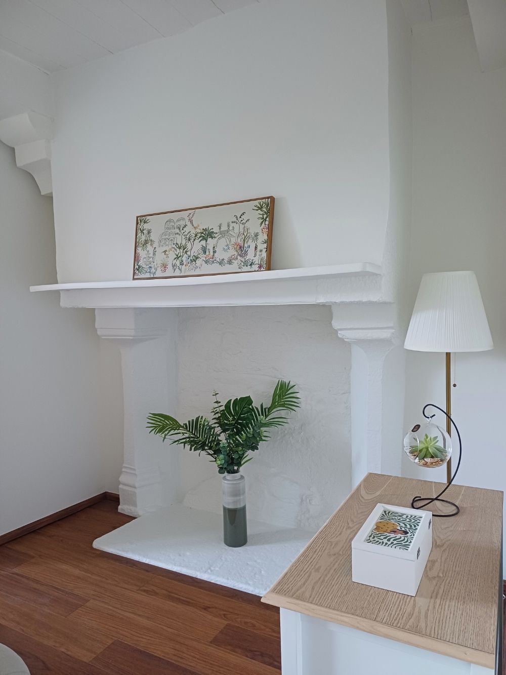 Een kamer met een witte open haard, een ingelijst kunstwerk erboven, een kamerplant, een lamp en een klein tafeltje met een boek.