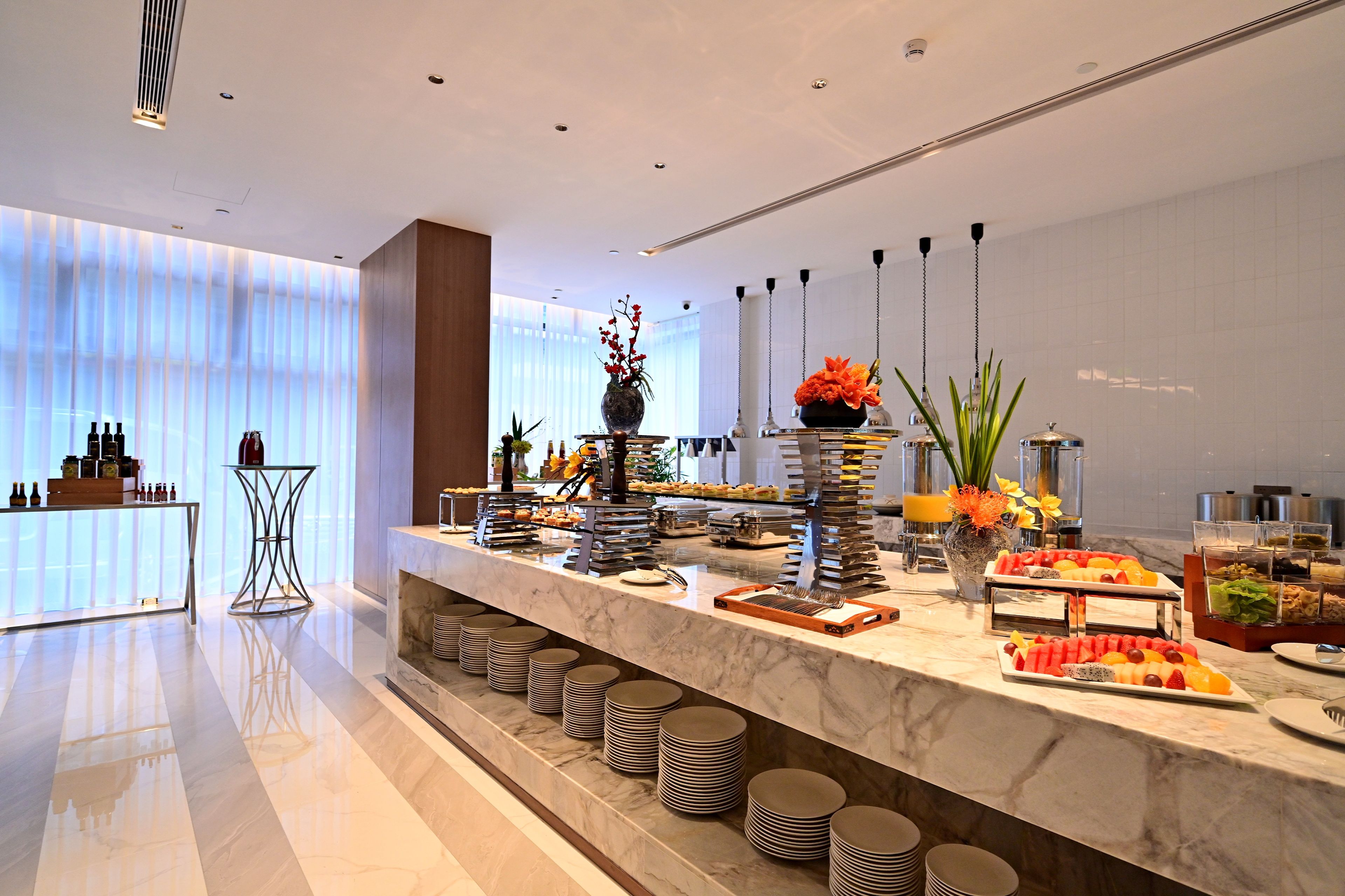 A modern hotel lobby with a marble reception desk, a glass-top table with bottles, and a buffet area with various food options.