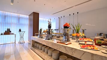 A modern hotel lobby with a marble reception desk, a glass-top table with bottles, and a buffet area with various food options.