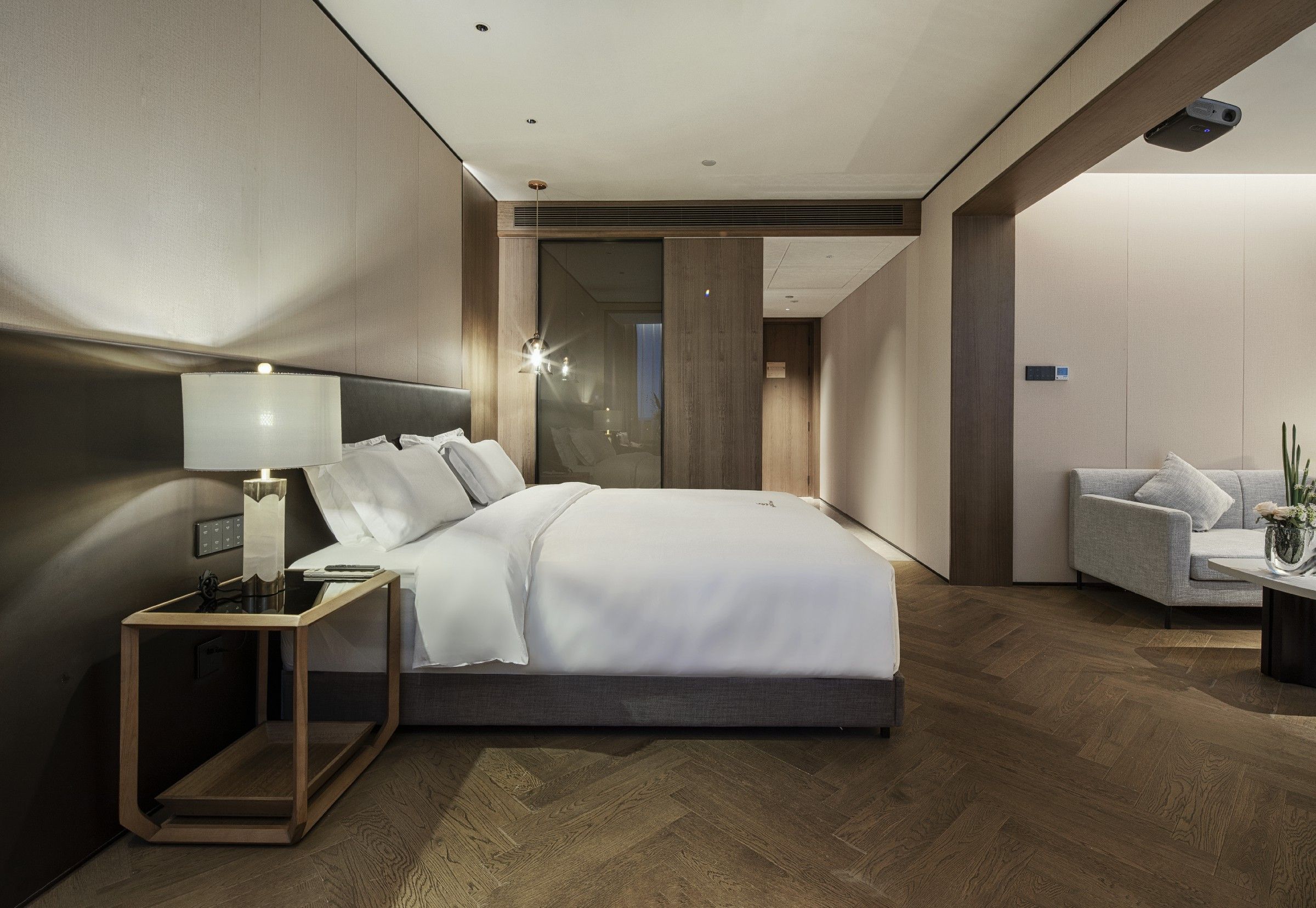 Una habitación de hotel moderna con una cama grande, mesita de noche, lámpara y una zona de estar con sofá y una pequeña mesa.