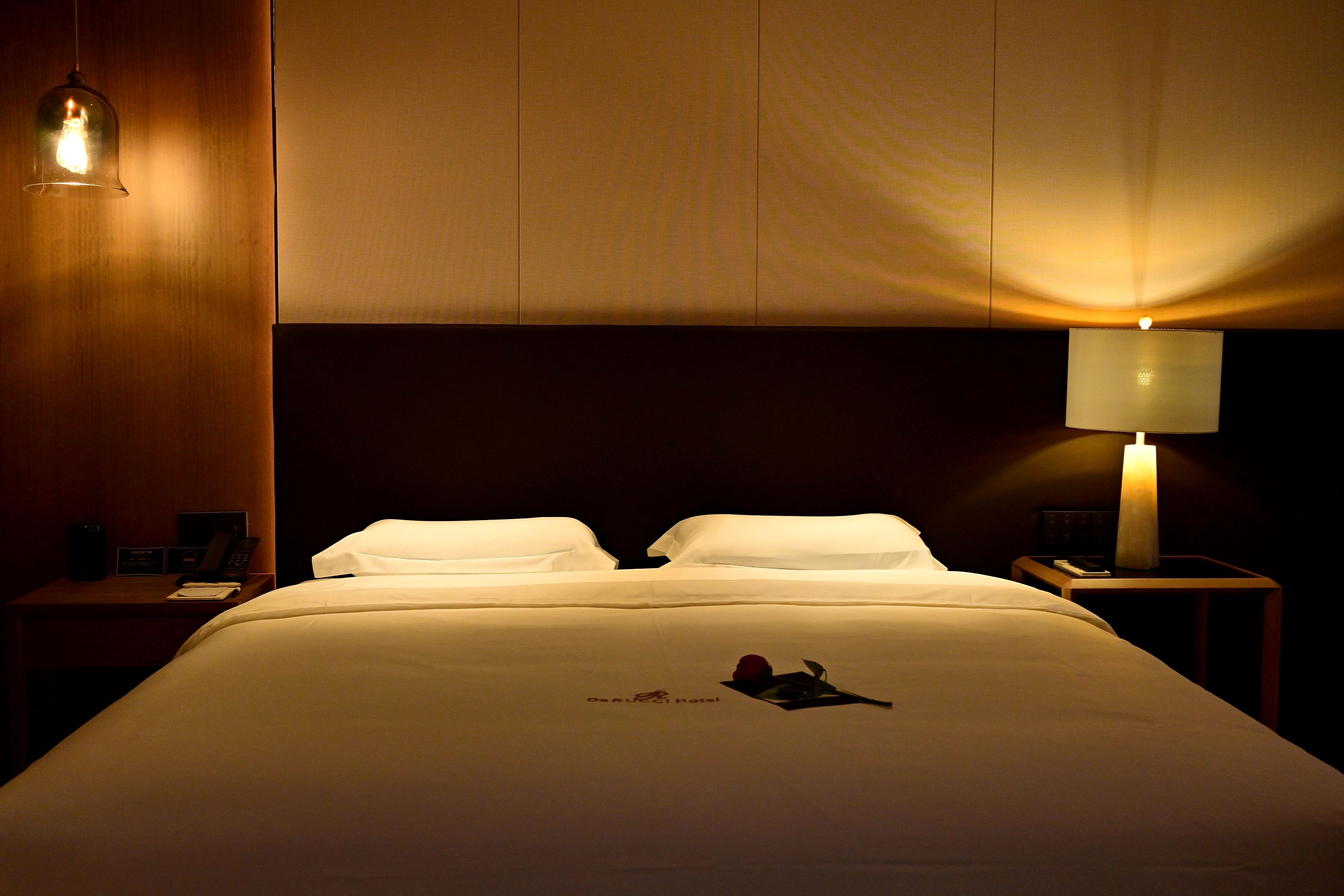 A hotel room with a bed, a nightstand, a lamp, and a hanging light fixture.