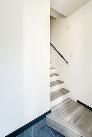 A staircase with wooden steps and a black handrail, leading to a landing with a white wall and a light switch. - HANASTAY MUKAI HOTARU (Osaka)