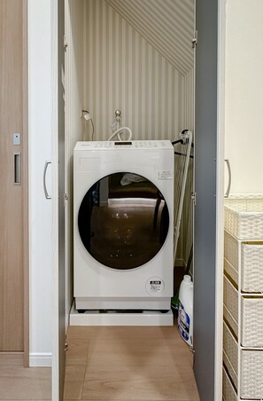 A compact, modern laundry room with a built-in washer and dryer, a small storage area, and a door leading to another room. - HANASTAY MUKAI HOTARU (Osaka)