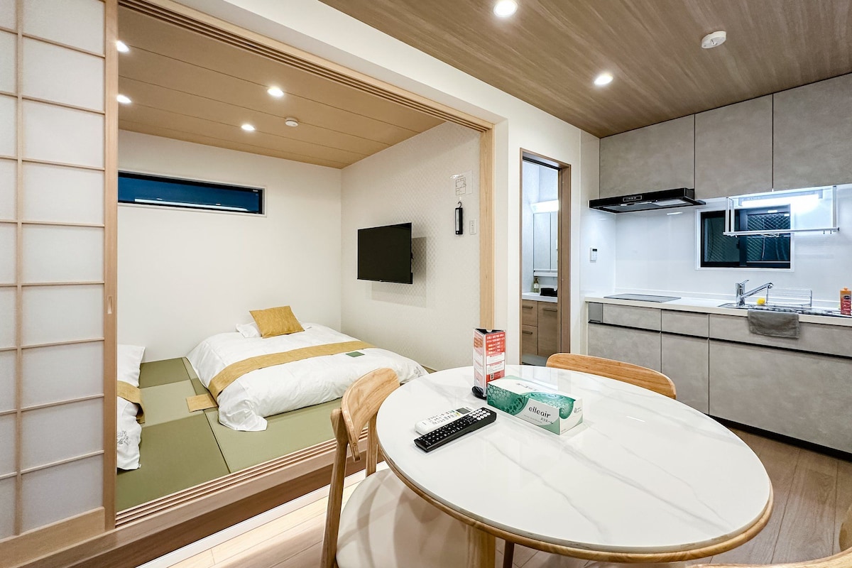 A modern hotel room with a bed, a dining table, a TV, and a kitchen area.