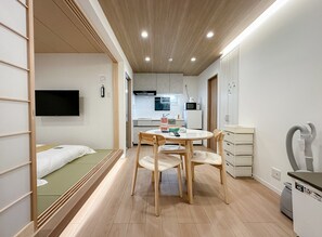 A compact hotel room with a bed, a small kitchenette, a dining area with a table and chairs, and a TV mounted on the wall. - HANASTAY MUKAI HOTARU (Osaka)