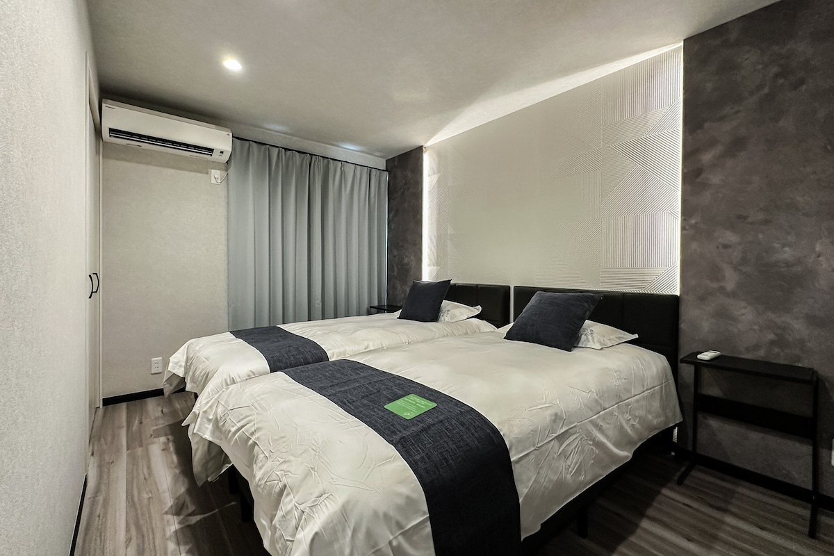A modern hotel room with a large bed, a bedside table, an air conditioning unit, and a window with blinds.