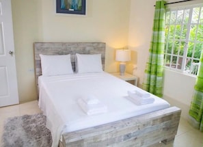 A neatly made bed with white linens and towels, a wooden headboard, a bedside lamp, and a window with green curtains. - A welcoming light open concept two bedroom apartment. (San Juan)