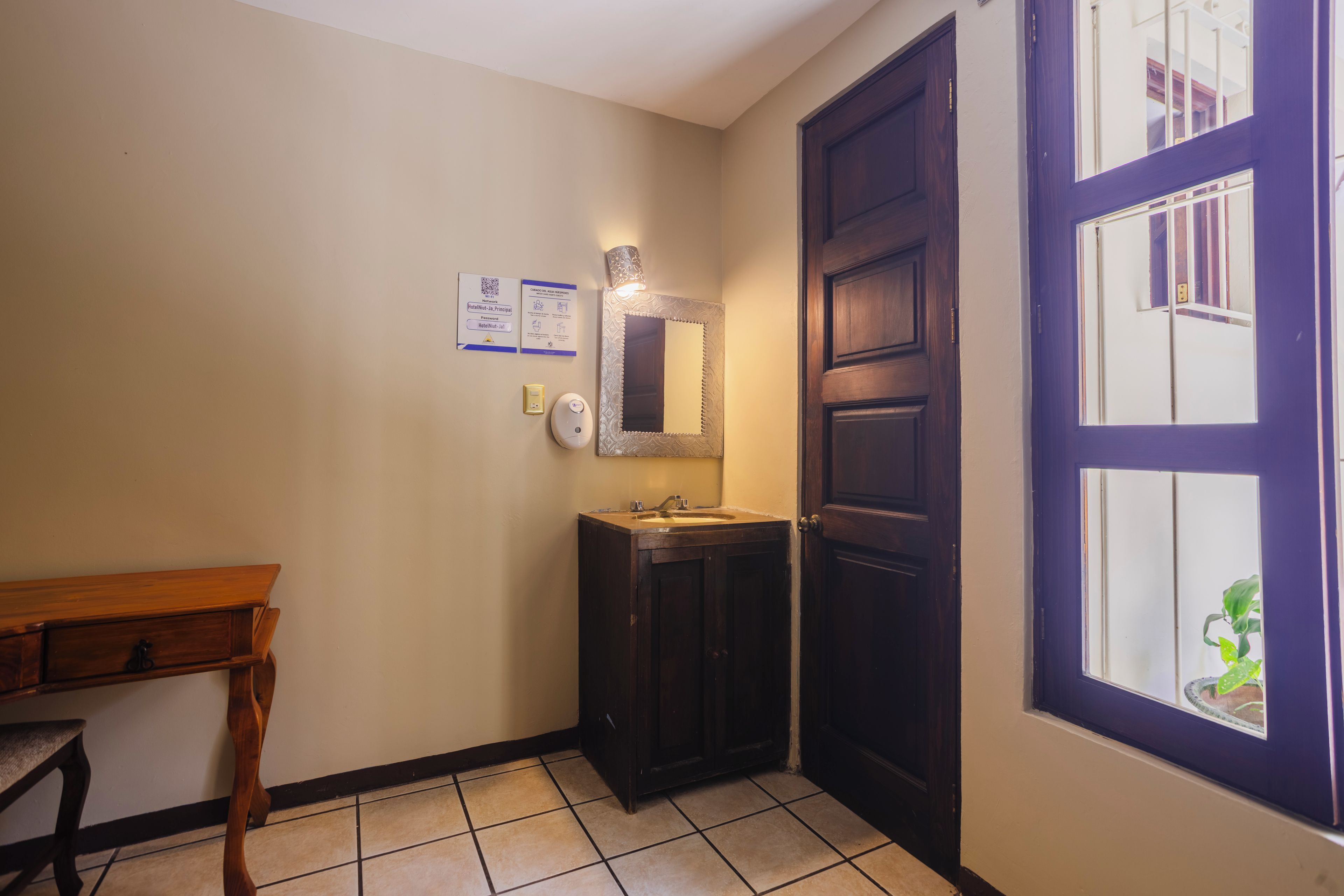 Un baño con lavamanos, espejo y una puerta con ventanal. Hay letreros en la pared y una mesita.