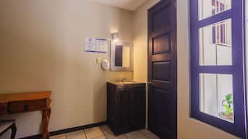 Un baño con lavamanos, espejo y una puerta con ventanal. Hay letreros en la pared y una mesita.