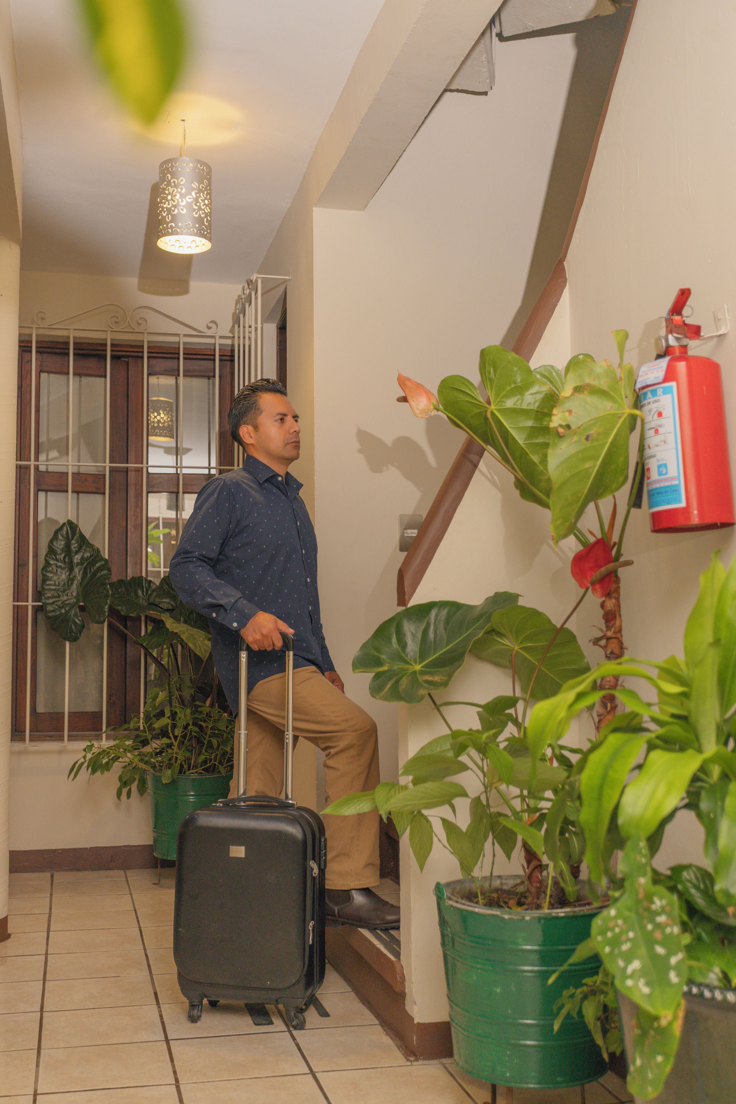 Una persona con una maleta en un pasillo con plantas en macetas y un extintor en la pared.