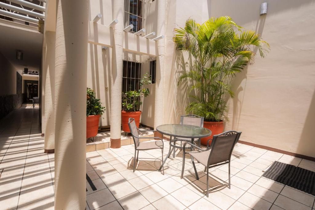 Un patio con una mesa y sillas de vidrio, rodeado de plantas en macetas y una pared blanca.