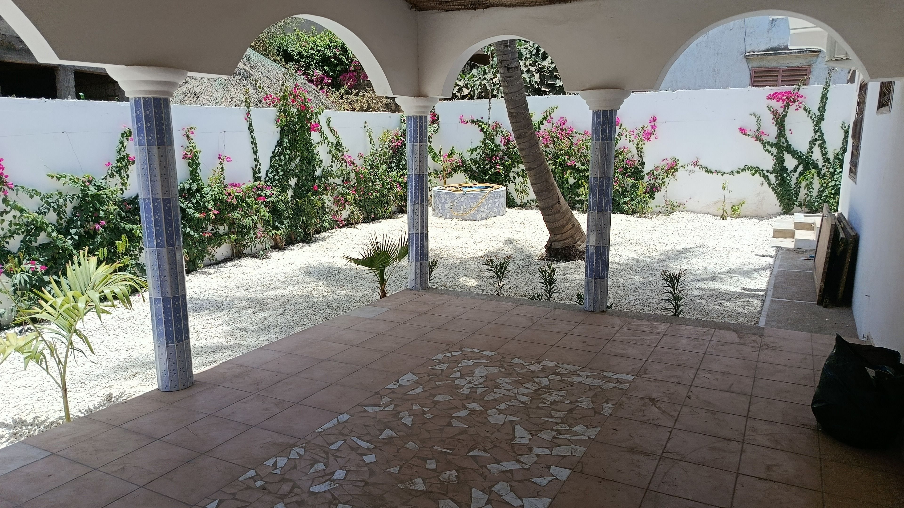 Un patio couvert doté d’un sol carrelé, de colonnes et d’un mur blanc orné de plantes à fleurs.