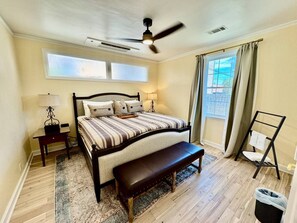 A bedroom with a bed, a bench, a nightstand, a lamp, a ceiling fan, a window with curtains, and a view of the outside. - Solost SOMA Little Rock (Little Rock)