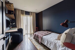 A bedroom with a bed, a desk, a chair, a red stool, and a window with curtains.