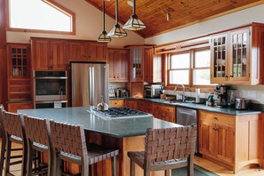 A kitchen with wooden cabinets, a central island with chairs, and a stove. - Cooperstown 5BR with Lake View, Hot Tub, Wiffle Ball Field on 15+ Private Acres (Richfield Springs)