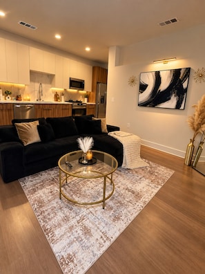 A modern living room with a black sofa, a glass coffee table, and a kitchen in the background. - The Luxe Noir near Downtown Atlanta (Atlanta)