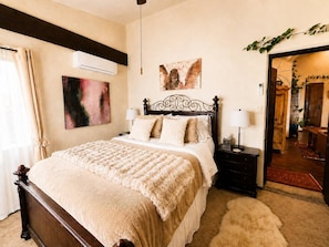 A bedroom with a large bed, a bedside table with a lamp, a wall-mounted air conditioner, and a painting above the bed.