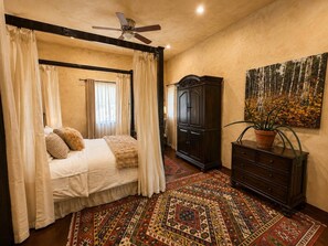 A bedroom with a four-poster bed, a dark wooden dresser, a large painting, and a patterned rug.