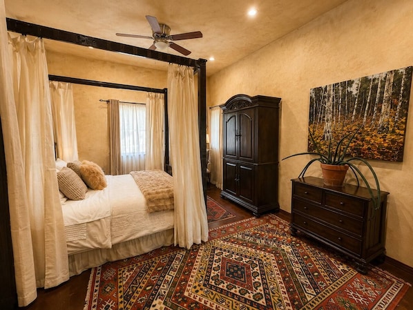 A bedroom with a four-poster bed, a dark wooden dresser, a large painting, and a patterned rug.
