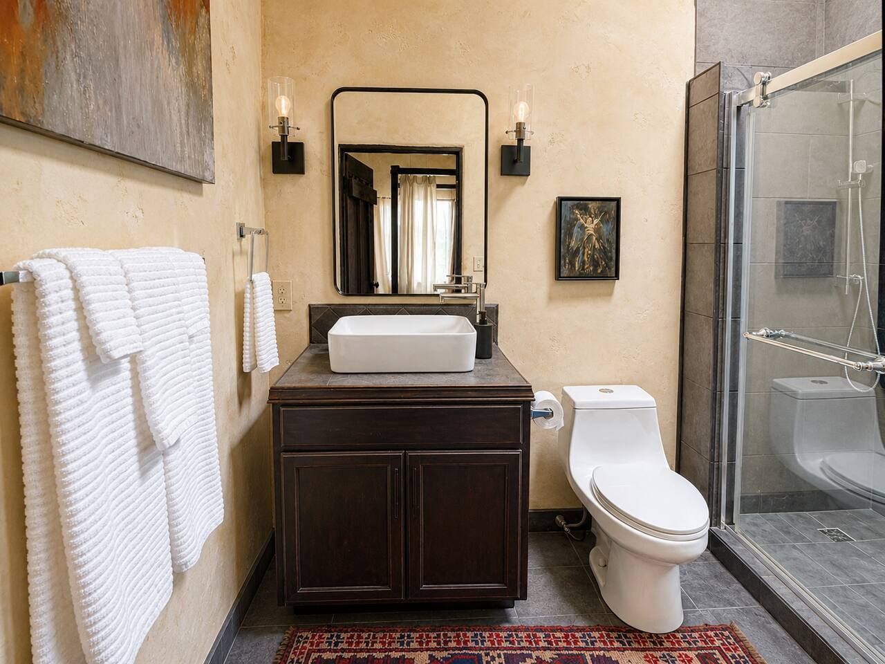A bathroom with a white sink, dark cabinets, a toilet, a shower, and a mirror with wall sconces.