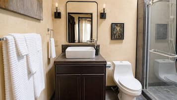 A bathroom with a white sink, dark cabinets, a toilet, a shower, and a mirror with wall sconces.