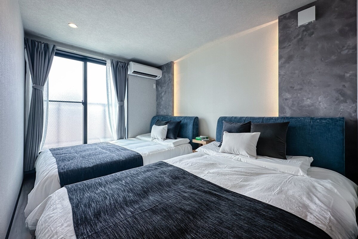 Ein Hotelzimmer mit zwei Betten, einem großen Fenster mit Vorhängen, einer Klimaanlage und einer strukturierten Wand.